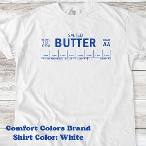 May include: White t-shirt with blue text and a butter measurement guide. The shirt features the words "SALTED BUTTER" and "GRADE AA". Measurements are in tablespoons and cups, with a net weight of 4 oz (113g). The shirt is a Comfort Colors brand.