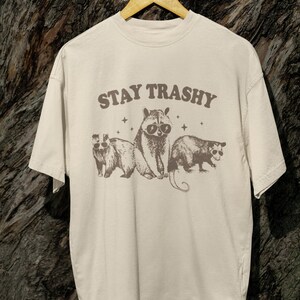 K&ouml;nnte beinhalten: Ein beiges T-Shirt mit einem grafischen Aufdruck von drei Tieren, die Sonnenbrillen tragen. Der Text "STAY TRASHY" ist in braunen Buchstaben &uuml;ber den Tieren gedruckt.