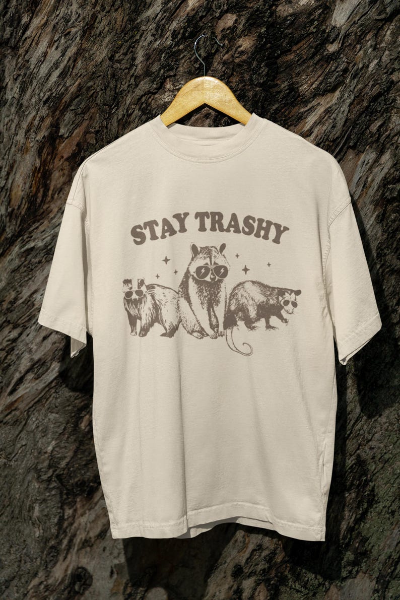 K&ouml;nnte beinhalten: Ein beiges T-Shirt mit einem grafischen Aufdruck von drei Tieren, die Sonnenbrillen tragen. Der Text "STAY TRASHY" ist in braunen Buchstaben &uuml;ber den Tieren gedruckt.