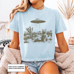 May include: Light blue chambray comfort colors cropped shirt featuring a graphic of a cat being abducted by a UFO in a desert landscape. The shirt has short sleeves and a crew neck.