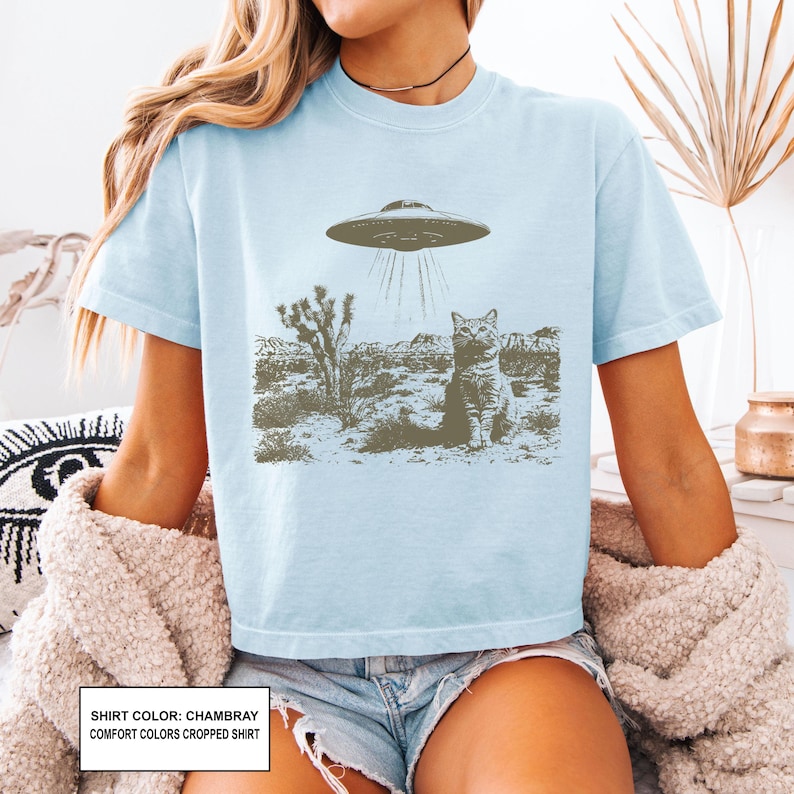 May include: Light blue chambray comfort colors cropped shirt featuring a graphic of a cat being abducted by a UFO in a desert landscape. The shirt has short sleeves and a crew neck.