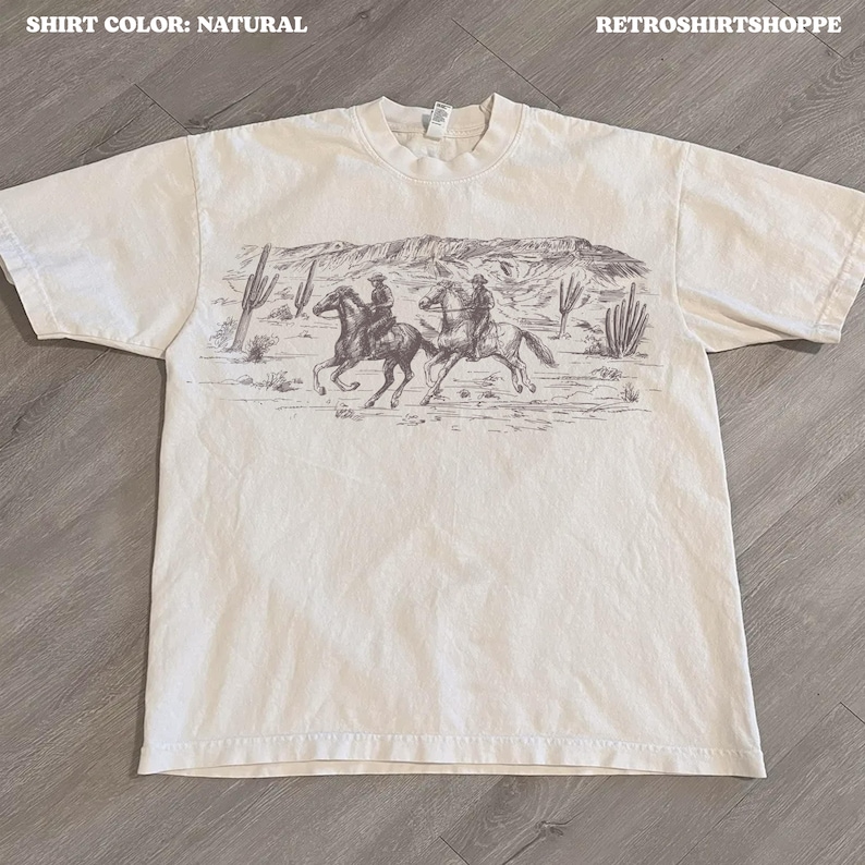 May include: Natural-colored t-shirt featuring a sepia-toned graphic of two cowboys riding horses through a desert landscape with cacti and mountains. The text "SHIRT COLOR: NATURAL" is at the top left, and "RETROSHIRTSHOPPE" is at the top right.