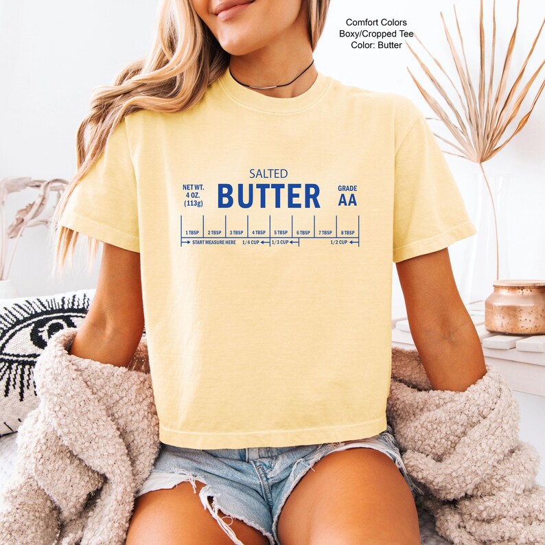 Puede incluir: Una camiseta amarilla Comfort Colors boxy/cropped con la palabra "BUTTER" en letras azules. La camiseta tiene un gr&aacute;fico de medidas de mantequilla, incluyendo "NET WT. 4 oz. (113g)" y "GRADE AA".