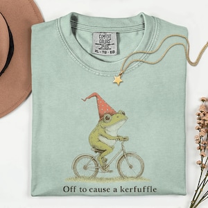 May include: A light green t-shirt featuring a whimsical illustration of a frog riding a bicycle and wearing a red hat. The text "Off to cause a kerfuffle" is printed below the image. A gold necklace with a star pendant is draped on the shirt.