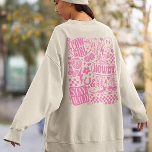 May include: Cream-colored sweatshirt with a pink graphic design on the back. The design includes the words "Cow Girl", "Howdy", and "Stay Wild", along with various cowboy-themed illustrations like boots, a horseshoe, and a cactus.