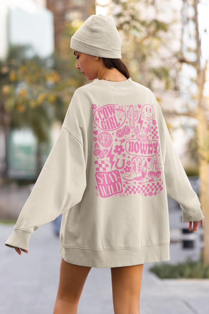 May include: Cream-colored sweatshirt with a pink graphic design on the back. The design includes the words "Cow Girl", "Howdy", and "Stay Wild", along with various cowboy-themed illustrations like boots, a horseshoe, and a cactus.