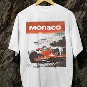 以下が含まれることがあります： 白いTシャツ。F1カーがトラックを走るレトロなグラフィック。車の上には赤いバナーで「Monaco」の文字。ヴィンテージ感のあるデザイン。