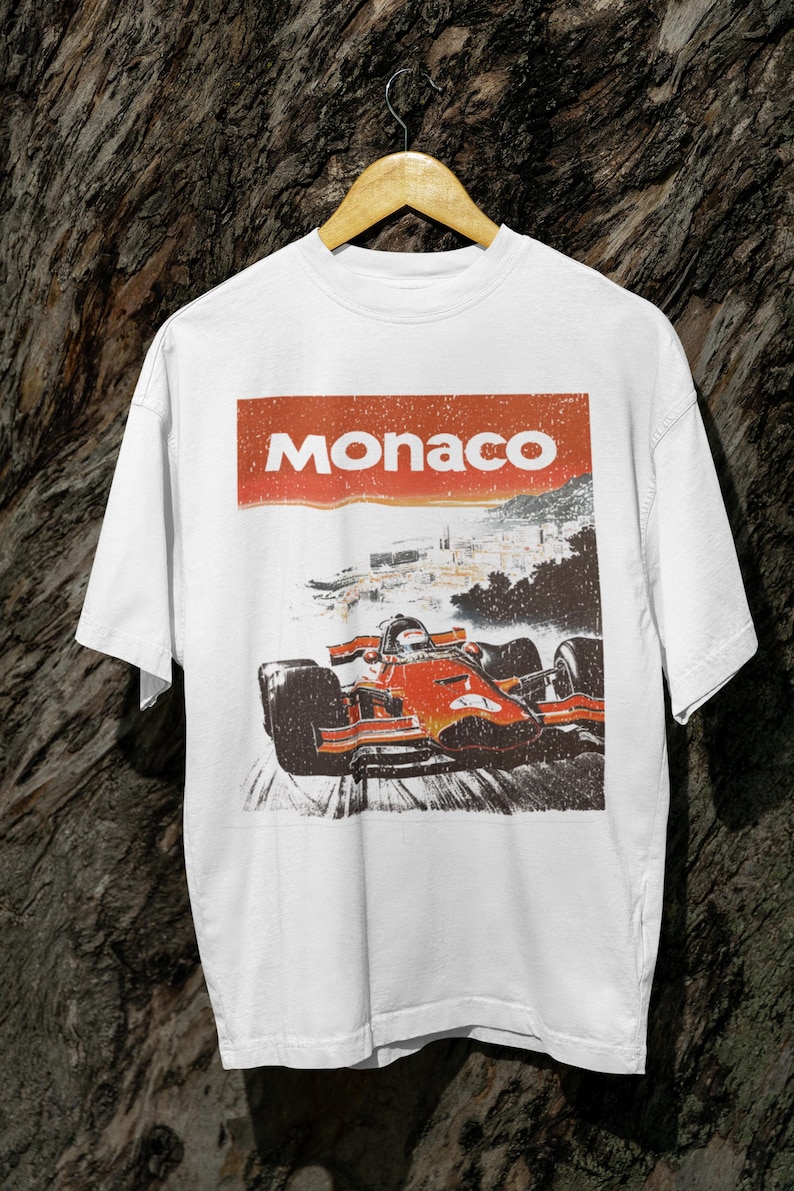 以下が含まれることがあります： 白いTシャツ。F1カーがトラックを走るレトロなグラフィック。車の上には赤いバナーで「Monaco」の文字。ヴィンテージ感のあるデザイン。