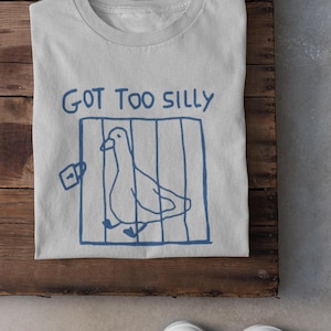 May include: A pale grey t-shirt featuring a blue drawing of a bird in a cage and the words "GOT TOO SILLY". The shirt is folded on a wooden surface, with white trainers visible at the bottom.