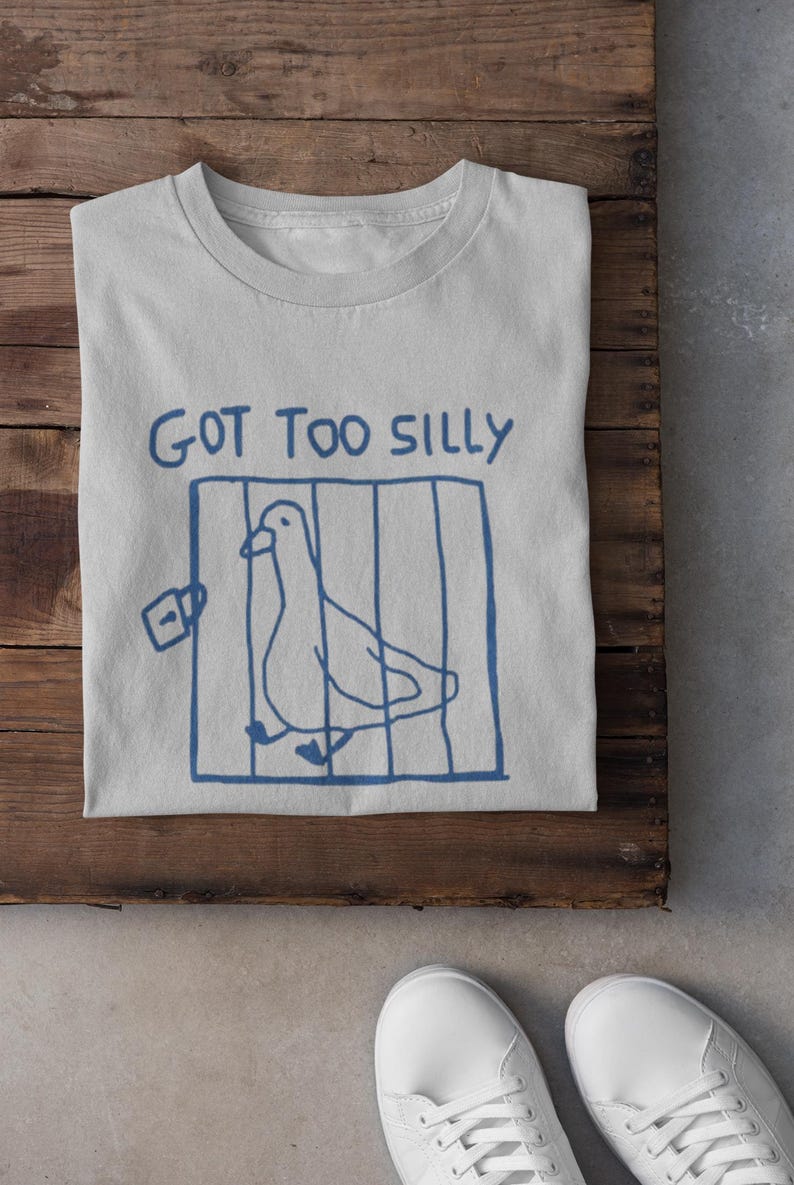 May include: A pale grey t-shirt featuring a blue drawing of a bird in a cage and the words "GOT TOO SILLY". The shirt is folded on a wooden surface, with white trainers visible at the bottom.