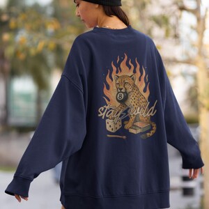 May include: Navy blue sweatshirt featuring a cheetah holding an 8-ball, set against a backdrop of flames. The phrase "stay wild" is written in a stylized font. A pair of dice and a matchbox are also included in the design.