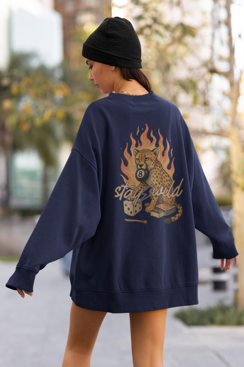 May include: Navy blue sweatshirt featuring a cheetah holding an 8-ball, set against a backdrop of flames. The phrase "stay wild" is written in a stylized font. A pair of dice and a matchbox are also included in the design.