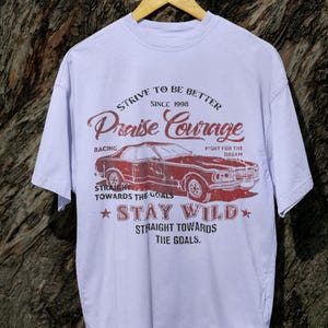 K&ouml;nnte beinhalten: Ein lavendelfarbenes T-Shirt mit einer Vintage-Grafik eines roten Autos. Die Grafik enth&auml;lt den Text "Strive to be better since 1998 Praise Courage Racing Fight for the Dream Straight towards the goals Stay Wild Straight towards the goals."