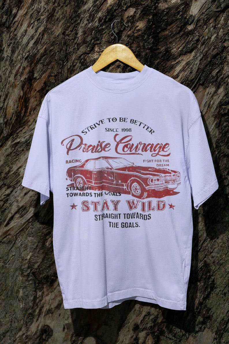 K&ouml;nnte beinhalten: Ein lavendelfarbenes T-Shirt mit einer Vintage-Grafik eines roten Autos. Die Grafik enth&auml;lt den Text "Strive to be better since 1998 Praise Courage Racing Fight for the Dream Straight towards the goals Stay Wild Straight towards the goals."