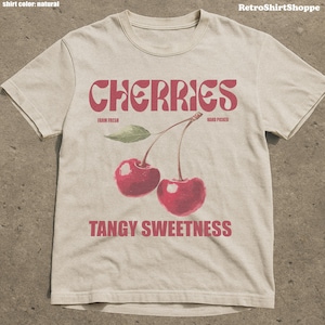 May include: A natural-colored t-shirt featuring a retro design with the word "CHERRIES" in a bold, red font. Below the word are two red cherries with green leaves and the words "FARM FRESH" and "HAND PICKED". The phrase "TANGY SWEETNESS" is printed below the cherries.