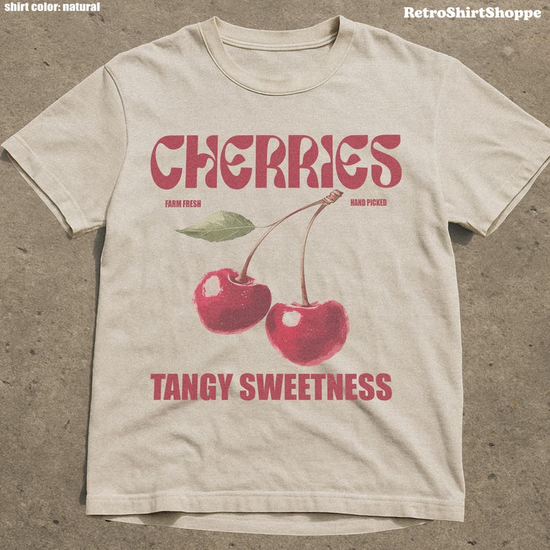 May include: A natural-colored t-shirt featuring a retro design with the word "CHERRIES" in a bold, red font. Below the word are two red cherries with green leaves and the words "FARM FRESH" and "HAND PICKED". The phrase "TANGY SWEETNESS" is printed below the cherries.