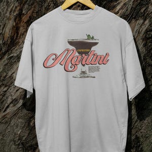 Puede incluir: Camiseta gris claro con un dise&ntilde;o de espresso martini. El dise&ntilde;o incluye las palabras "Espresso Martini" en una fuente retro, con una ilustraci&oacute;n de copa de c&oacute;ctel. La camiseta est&aacute; colgada en una percha de madera.