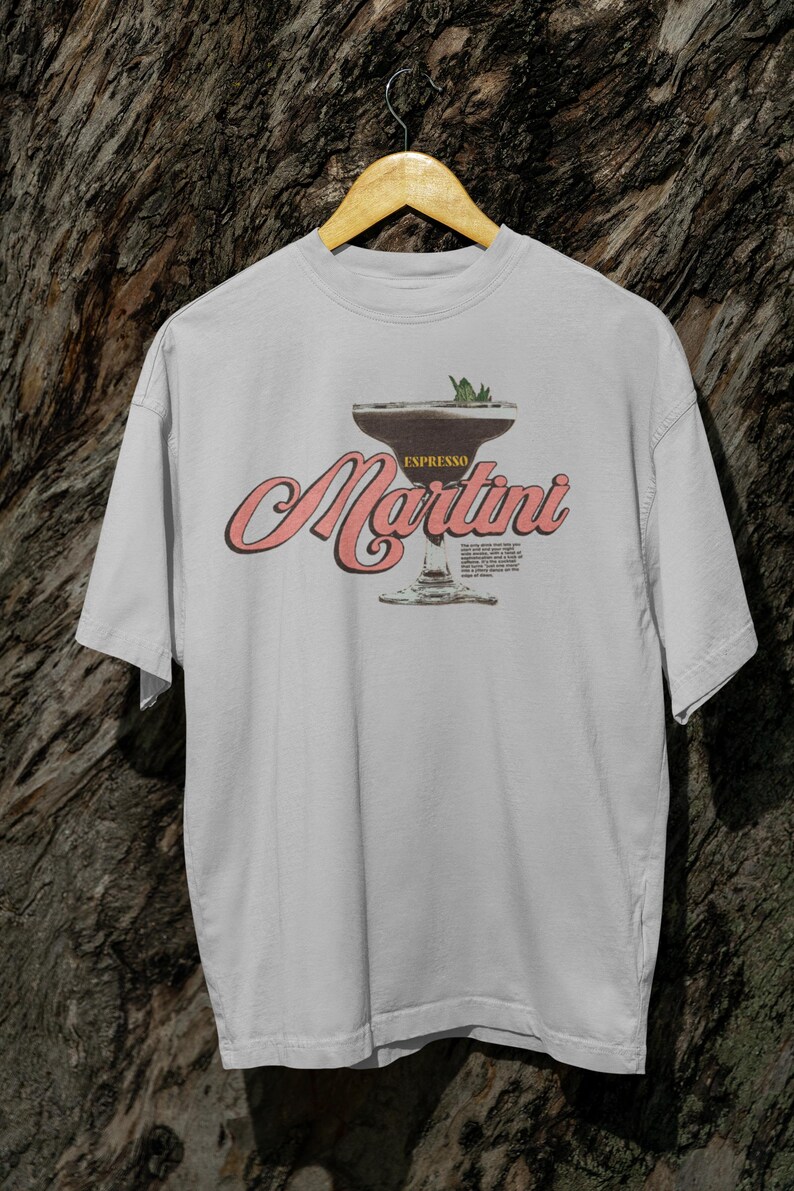 Puede incluir: Camiseta gris claro con un dise&ntilde;o de espresso martini. El dise&ntilde;o incluye las palabras "Espresso Martini" en una fuente retro, con una ilustraci&oacute;n de copa de c&oacute;ctel. La camiseta est&aacute; colgada en una percha de madera.