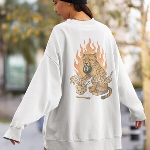 May include: White sweatshirt with a graphic of a cheetah holding a pool ball with the number 8, flames, dice, and the words "Stay Wild". The sweatshirt is a relaxed fit.