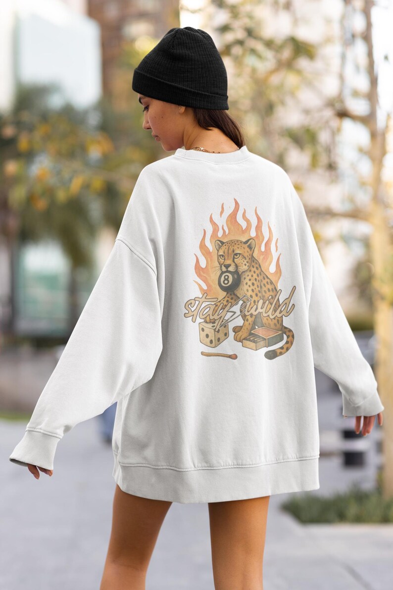 May include: White sweatshirt with a graphic of a cheetah holding a pool ball with the number 8, flames, dice, and the words "Stay Wild". The sweatshirt is a relaxed fit.