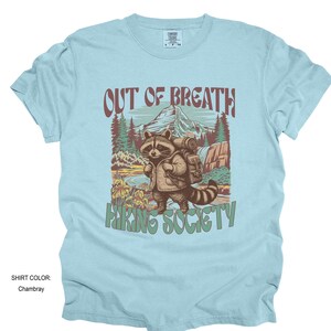 May include: Light blue t-shirt with a graphic of a raccoon wearing a backpack, standing in a mountain landscape. Text reads "OUT OF BREATH" above and "HIKING SOCIETY" below. The shirt color is Chambray.