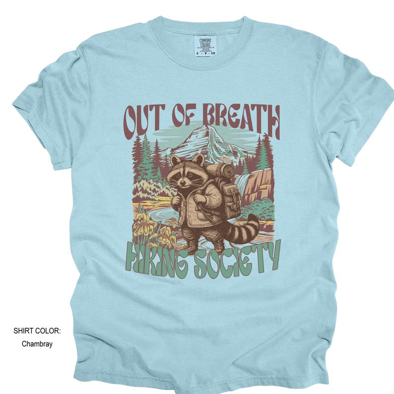 May include: Light blue t-shirt with a graphic of a raccoon wearing a backpack, standing in a mountain landscape. Text reads "OUT OF BREATH" above and "HIKING SOCIETY" below. The shirt color is Chambray.