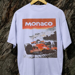 以下が含まれることがあります： オレンジと赤のフォーミュラ1レーシングカーのグラフィックが特徴の白いTシャツ。上部には赤い長方形に「Monaco」の文字。 ヴィンテージ感のあるデザイン。