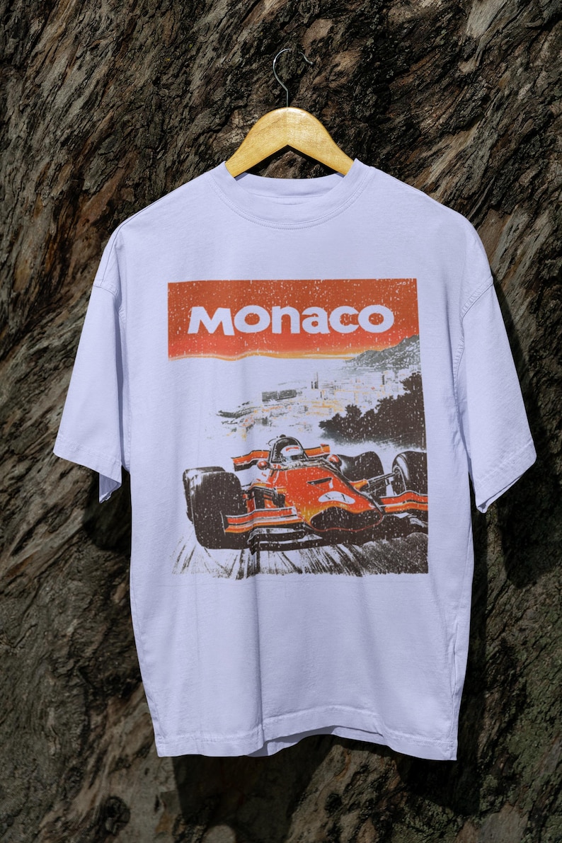 以下が含まれることがあります： オレンジと赤のフォーミュラ1レーシングカーのグラフィックが特徴の白いTシャツ。上部には赤い長方形に「Monaco」の文字。 ヴィンテージ感のあるデザイン。