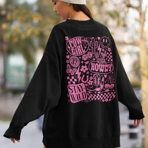 May include: Black sweatshirt with a pink graphic design featuring the words "Cow Girl", "Howdy", and "Stay Wild". The design includes a smiley face, cactus, boots, and other western-themed elements. The sweatshirt is a casual style.