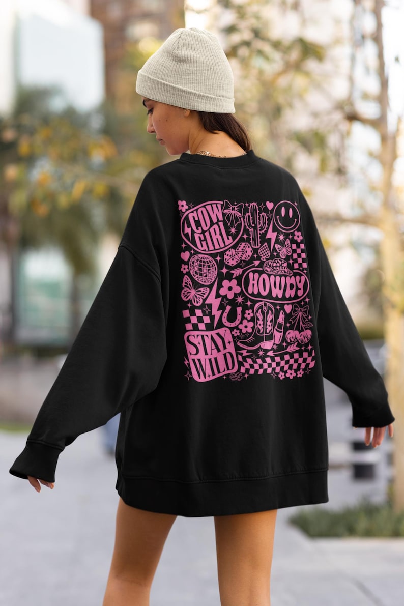 May include: Black sweatshirt with a pink graphic design featuring the words "Cow Girl", "Howdy", and "Stay Wild". The design includes a smiley face, cactus, boots, and other western-themed elements. The sweatshirt is a casual style.