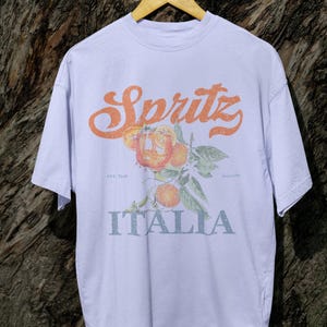 May include: A light purple t-shirt with the word "Spritz" in orange script above a graphic of oranges and leaves. Below the graphic, the word "ITALIA" is printed in a light blue color. The shirt is hanging on a wooden hanger.