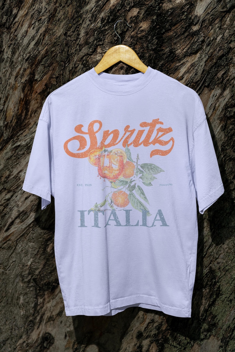 May include: A light purple t-shirt with the word "Spritz" in orange script above a graphic of oranges and leaves. Below the graphic, the word "ITALIA" is printed in a light blue color. The shirt is hanging on a wooden hanger.