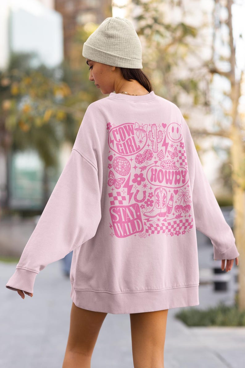 May include: A light pink sweatshirt with a graphic design on the back. The design includes the words "Cow Girl", "Howdy", and "Stay Wild", along with various illustrations such as a cactus, butterflies, and boots.