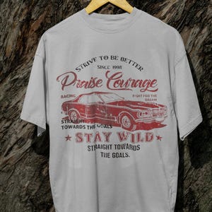 K&ouml;nnte beinhalten: Ein graues T-Shirt mit einem roten Vintage-Grafikdesign, das ein Auto, den Text "Strive to be better since 1998 Praise Courage" und den Text "Straight towards the goals Stay Wild Straight towards the goals." zeigt.