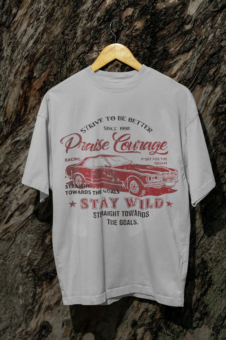 K&ouml;nnte beinhalten: Ein graues T-Shirt mit einem roten Vintage-Grafikdesign, das ein Auto, den Text "Strive to be better since 1998 Praise Courage" und den Text "Straight towards the goals Stay Wild Straight towards the goals." zeigt.