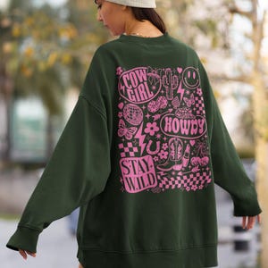 May include: Dark green sweatshirt with a pink graphic design on the back. The design includes the words "Cow Girl", "Howdy", and "Stay Wild", along with various illustrations such as a cactus, butterflies, and a horseshoe.