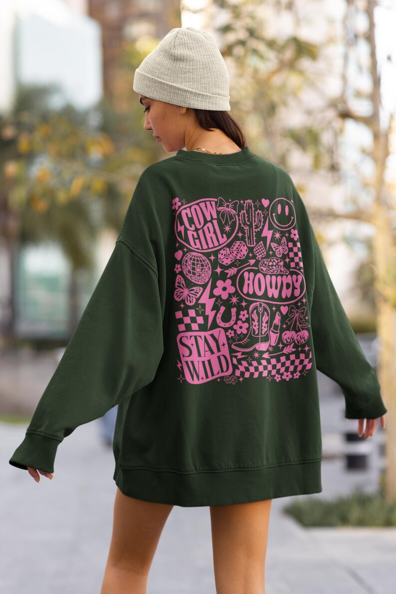 May include: Dark green sweatshirt with a pink graphic design on the back. The design includes the words "Cow Girl", "Howdy", and "Stay Wild", along with various illustrations such as a cactus, butterflies, and a horseshoe.