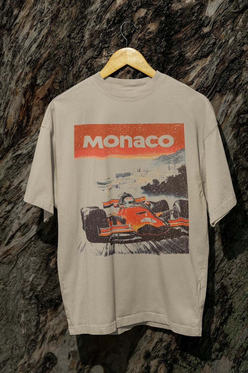 以下が含まれることがあります： ベージュ色のTシャツ。レトロなグラフィックで、フォーミュラ1レーシングカーが描かれています。「Monaco」の文字がオレンジ色の長方形で車の真上にプリントされています。木製のハンガーにかけられています。