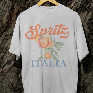 May include: White t-shirt with a vintage-style design featuring the word "Spritz" in orange, a branch of oranges, and the word "Italia" in blue. The shirt has a relaxed fit and short sleeves.