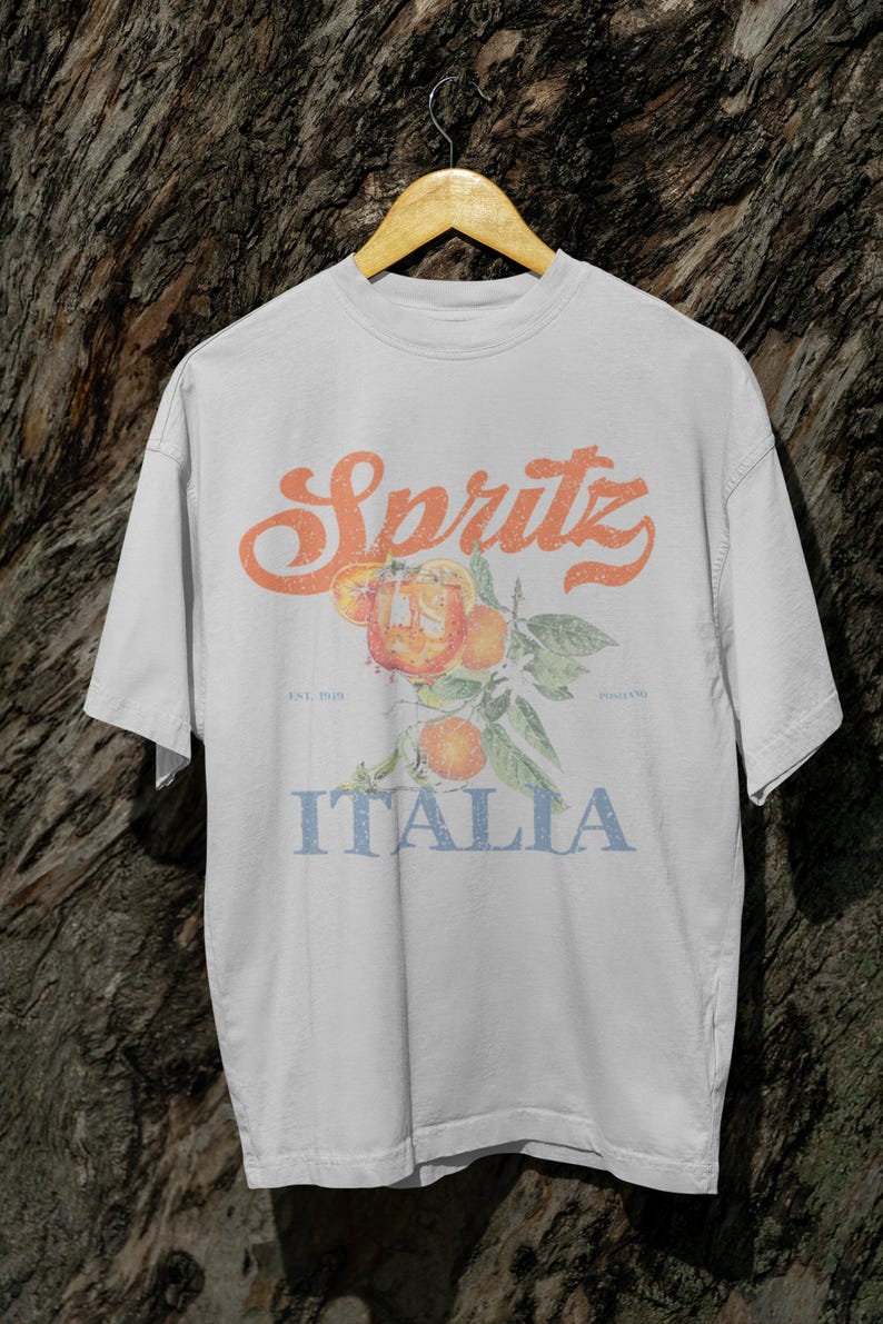 May include: White t-shirt with a vintage-style design featuring the word "Spritz" in orange, a branch of oranges, and the word "Italia" in blue. The shirt has a relaxed fit and short sleeves.