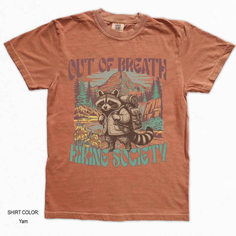 May include: Yam-colored t-shirt with a graphic of a raccoon wearing a backpack, standing in a mountain landscape. Text on the shirt reads "OUT OF BREATH" and "HIKING SOCIETY".