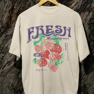 May include: A beige t-shirt with a graphic print of raspberries and the text "FRESH Raspberries Enjoy Little Things Sweet Fresh And Tart".