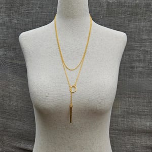 May include: A gold layered necklace with a long pendant featuring a circle and a vertical bar.