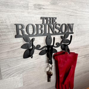 Può includere: Portachiavi e attaccapanni da parete in metallo nero con il nome "THE ROBINSON" ritagliato. Il portachiavi presenta decorazioni a forma di foglie e ganci. Un paio di chiavi e un guanto rosso sono appesi ai ganci.