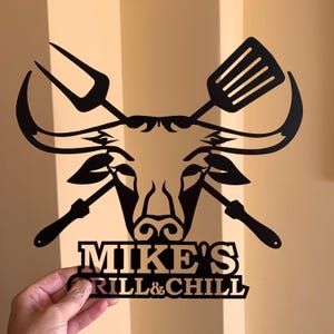 Black metal wall art featuring a bull's head with crossed grilling tools and the text "MIKE'S GRILL & CHILL".