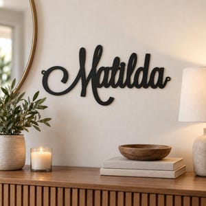 May include: A black metal wall sign featuring the name Matilda in a cursive script font. The sign has a decorative swirl on the left side and small circular mounting holes at both ends, displaying the name in a bold, dark, matte finish.