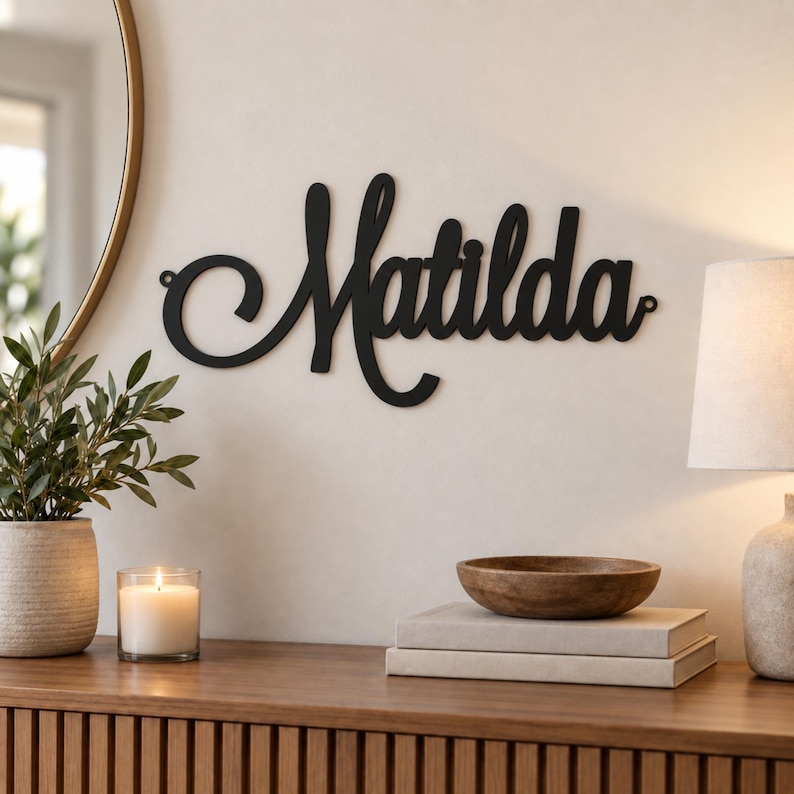 May include: A black metal wall sign featuring the name Matilda in a cursive script font. The sign has a decorative swirl on the left side and small circular mounting holes at both ends, displaying the name in a bold, dark, matte finish.