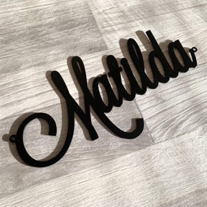 May include: A black wall sign featuring the name Matilda written in a continuous cursive script font. The sign is constructed from a flat material and includes small circular mounting holes at both the far left and far right ends of the lettering.