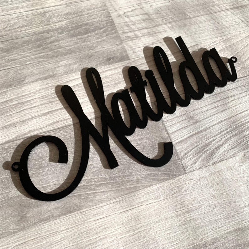 May include: A black wall sign featuring the name Matilda written in a continuous cursive script font. The sign is constructed from a flat material and includes small circular mounting holes at both the far left and far right ends of the lettering.