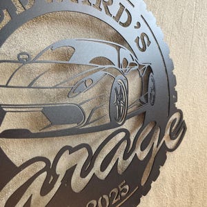 Op de afbeelding: Zwart metalen bord met een silhouet van een sportwagen en de tekst "[Name]'s Garage" met het jaar 2025 eronder.
